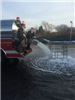 Two Firefighters on the Back of a Fire Vehicle with Water Pouring out into a Body of Water