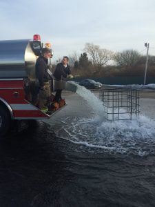 Two Firefighters on the Back of a Fire Vehicle with Water Pouring out into a Body of Water